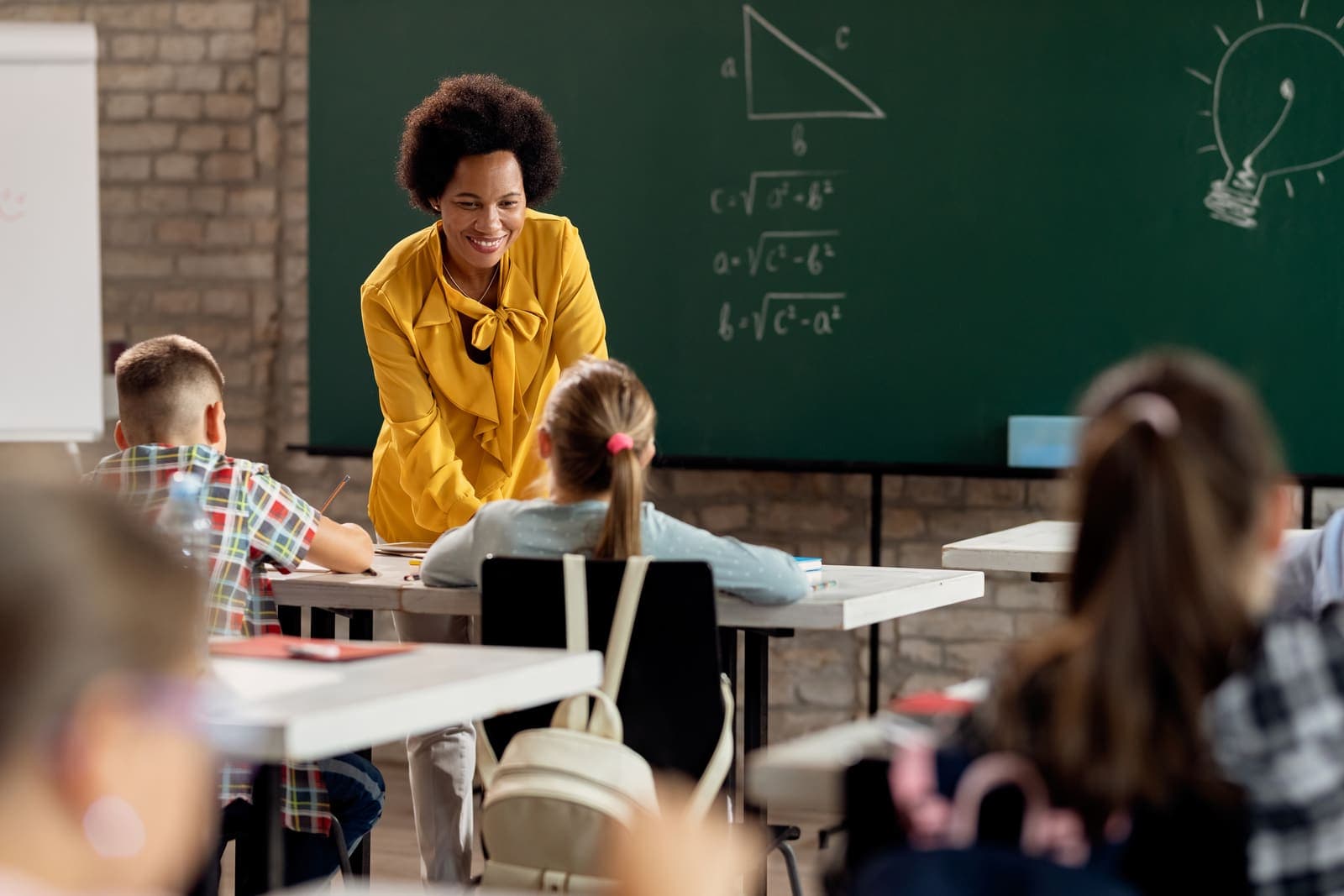 Professora conversando com alunos em uma sala de aula, com um quadro-negro cheio de fórmulas ao fundo