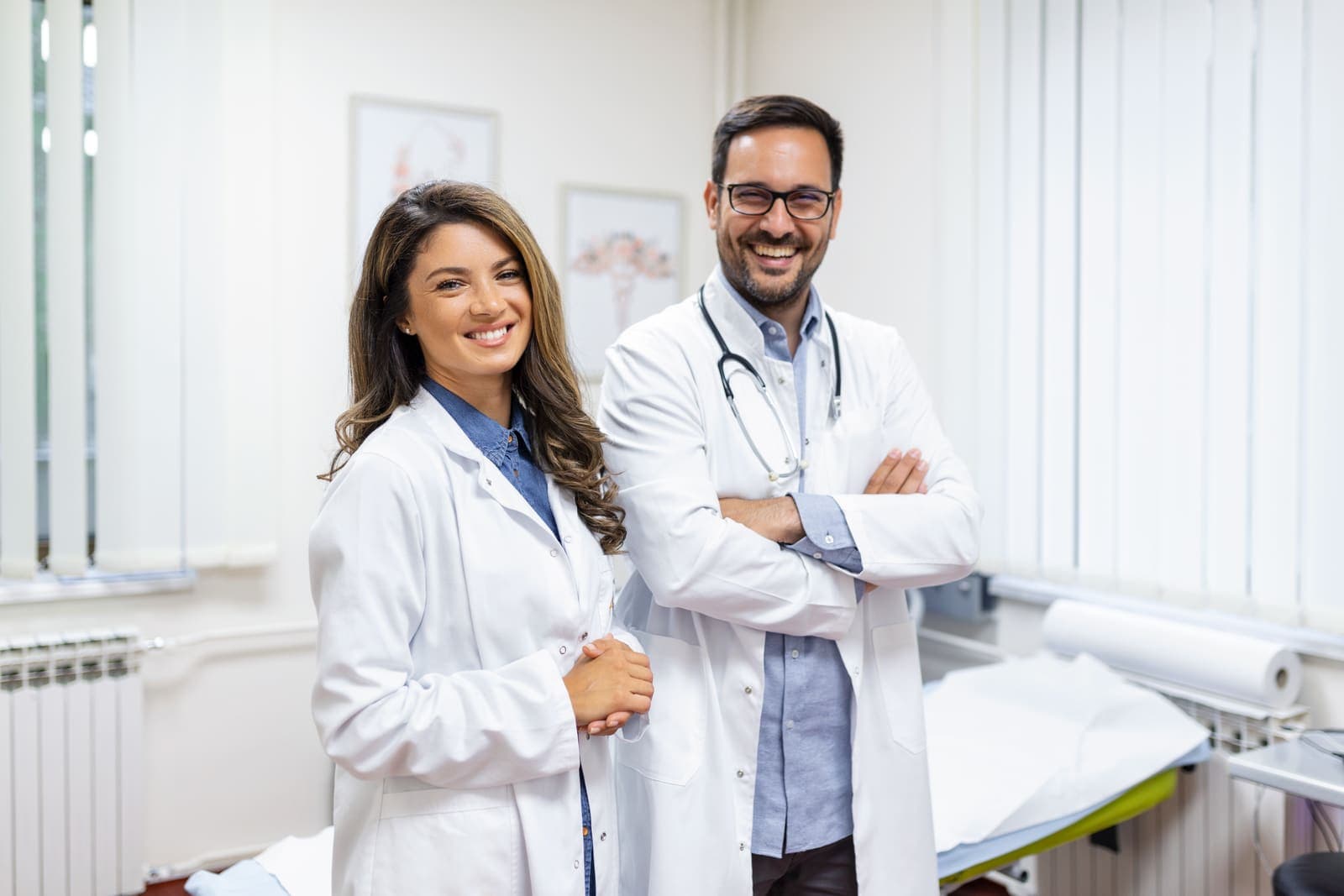 Uma médica e um médico de jaleco, sorrindo em um consultório